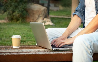 Genç adam halka açık bir parkta otururken dizüstü bilgisayarıyla çalışıyor ve kahve içiyor.