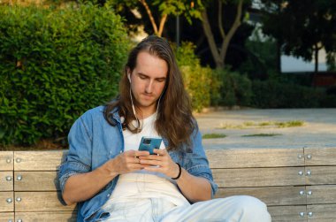 Genç adam parkta otururken cep telefonundan video izliyor.
