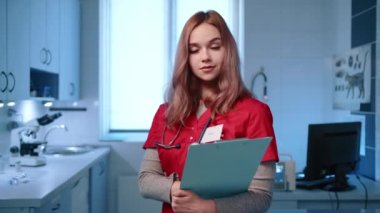 Kameraya gülümseyen ve poz veren genç bayan veterinerlere yakın çekim. Doktor kırmızı doktor kıyafeti giyiyor ve reçeteli şablon tutuyor. Yüksek kalite 4k görüntü