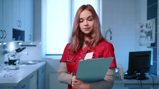 身边的年轻美丽的女兽医笑着对着镜头摆姿势 医生穿着红色的医生服 拿着处方模板 高质量的4K镜头 — 图库视频影像