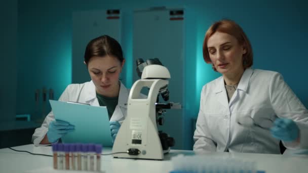 Kvinnlig Forskare Tittar Ett Mikroskop Medan Annan Skriver Ner Något — Stockvideo