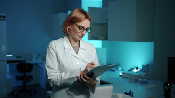 一个戴着眼镜 穿着白色实验室外套的兽医在石碑上写着什么 面带微笑 她在实验室里 戴着眼镜 她很专注 — 图库视频影像