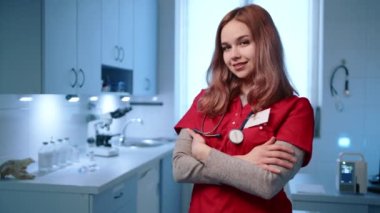 Gülümseyen genç bayan veteriner, hayvan hastanesinin açık modern ofisinde duruyor. Doktor steteskoplu doktor kıyafeti giyiyor, kolları çapraz. Yüksek kalite 4k görüntü