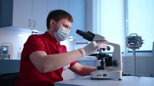 年轻的男性严肃的动物学家用双目显微镜检查样本进行分析 兽医有红色的医生服 乳胶手套和医疗面罩 高质量的4K镜头 — 图库视频影像