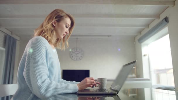 迷人的女人在家里的笔记本电脑上输入信息 金发姑娘专注于工作 杯子立在笔记本电脑旁边 后面有灯光室高质量的4K镜头 — 图库视频影像