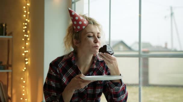 一个戴着派对帽的伤心女人坐在窗前吃一块巧克力蛋糕 她脸上有流口水的妆容 看起来很沮丧高质量的4K镜头 — 图库视频影像