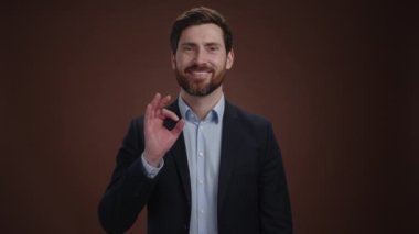 Takım elbiseli bir adam tamam işareti yapıyor ve kameraya gülümsüyor. Güzel giyinmiş ve çok mutlu görünüyor. O, kahverengi bir arka plandadır. Yüksek kalite 4k görüntü