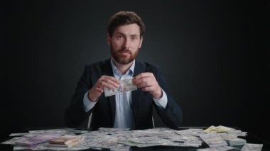 Takım elbiseli bir adam kameraya bakıyor ve banknotu ikiye bölüyor. Kendini beğenmişe benziyor. Adam siyah odada yatıyor. Para büyük bir yığının içinde. Yüksek kalite 4K görüntü. 