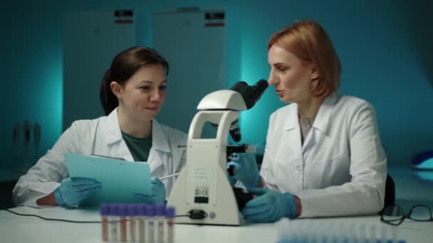 Kvinnlig Forskare Tittar Igenom Ett Mikroskop Medan Annan Skriver Något — Stockvideo