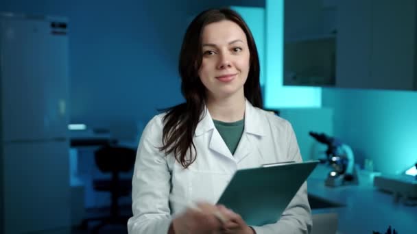 一位女科学家一边拿着平板电脑一边笑着看着摄像机 她在一个蓝色灯火通明的实验室里 看上去很友善 高质量的4K镜头 — 图库视频影像