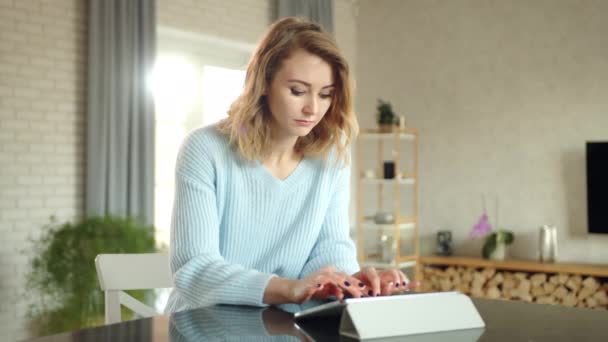 年轻漂亮的女人穿着白衣在平板电脑上打字想了一会儿 穿着蓝色毛衣的女人坐在舒适公寓的办公桌前 桌子上的石碑高质量的4K镜头 — 图库视频影像