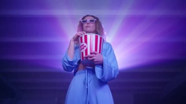 Beyaz kadın 3 boyutlu gözlüklü bir film izliyor ve evde patlamış mısır yiyor. Kıvırcık sarı saçlı, heyecanlı genç bir bayana yaklaş. Arka planda projektör ışınları var. Yüksek kalite 4k görüntü