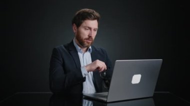 Siyah takım elbiseli yorgun bir adam gözlük takıyor ve dizüstü bilgisayarıyla çalışmaya devam ediyor. Gerçekten odaklanmış görünüyor. O, siyah bir odada. Yüksek kalite 4k görüntü