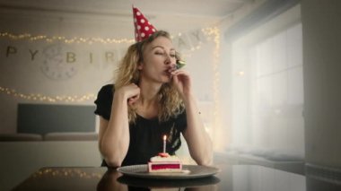 Parti şapkalı bir kadın doğum günü pipisini üflüyor ve mum çırpıyor. Bir kadın açık bej bir odada, süslemeleri ve küçük bir pasta dilimi ile. yüksek kalite 4k görüntü