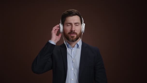 スーツを着た男とヘッドフォンで音楽を聴きながらヘッドセットを手にしている男 彼は単純な暗褐色の背景をしている 彼は驚いているようだ 高品質4K映像 — ストック動画