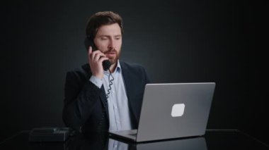 Siyah takım elbiseli bir adam laptopuyla çalışıyor ve masadaki iş telefonuna cevap veriyor. O, işine odaklanmış görünüyor. O, siyah bir odada. Yüksek kalite 4k görüntü 