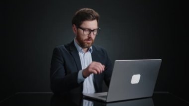 Siyah takım elbiseli bir adam dizüstü bilgisayarı üzerinde çalışırken gözlüğünü çıkarıyor ve gözlerini siliyor. Çok mutlu görünüyor. O, siyah bir odada. Yüksek kalite 4k görüntü