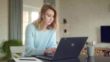 Bilgileri araştırmaya odaklanmış, dizüstü bilgisayardaki tipler. Kadın evinde rahat bir dairede çalışıyor. Laptop, kağıt, bardak masanın üstünde. Yüksek kalite 4k görüntü