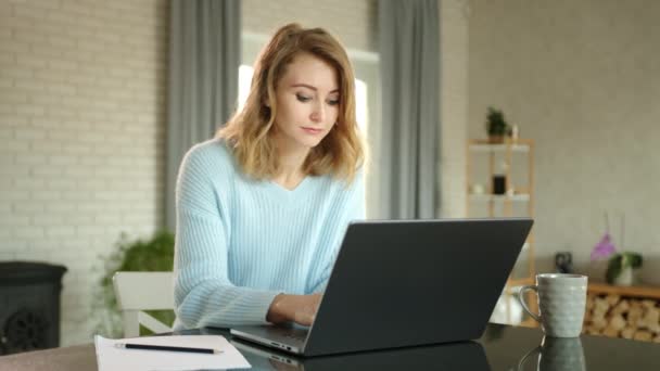 迷人的微笑的年轻女子在家里舒适的灯光下在笔记本电脑上打字 金发姑娘在笔记本电脑旁边有一杯茶和一张铅笔纸 高质量的4K镜头 — 图库视频影像