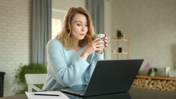 可爱的金发美女喝茶 并通过笔记本电脑上的视频链接进行对话 女人手里拿着杯子 坐在桌旁 后面有一个舒适的房间 高质量的4K镜头 — 图库视频影像