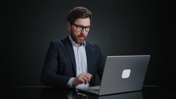 一名身穿黑色西服 戴着眼镜的男子一边在笔记本电脑前微笑 一边在笔记本电脑上工作 他看起来真的很高兴 他在一间黑屋里 高质量的4K镜头 — 图库视频影像