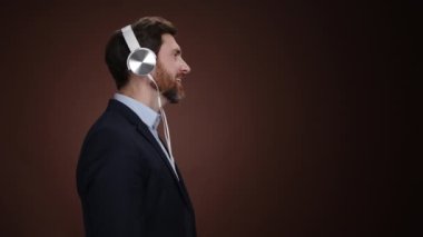Takım elbiseli ve kulaklıklı bir adam kafasını sallıyor ve biraz dans ediyor. O, basit bir koyu kahverengi arka plandadır. Şaşırmış görünüyor. Yüksek kalite 4k görüntü
