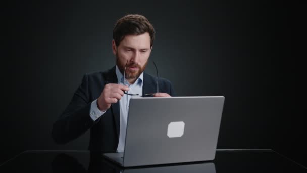 眼鏡をかけた黒いスーツの男がノートパソコンで働き始めた 彼は仕事に集中しているようだ 彼は黒い部屋にいる 高品質4K映像 — ストック動画