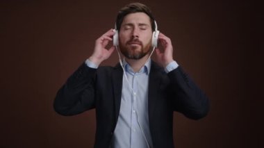 Takım elbiseli ve kulaklıklı bir adam gözleri kapalı müzik dinliyor. O, basit bir koyu kahverengi arka plandadır. Şaşırmış görünüyor. Yüksek kalite 4k görüntü