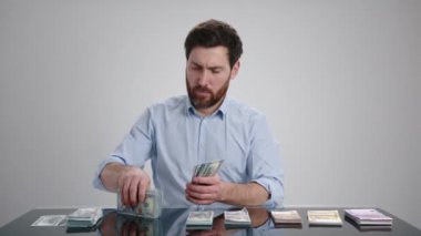 Mavi gömlekli bir adam para sayıyor ve masanın üzerine yığıyor. Odaklanmış görünüyor. Adam açık gri bir odada. Yüksek kalite 4k görüntü 