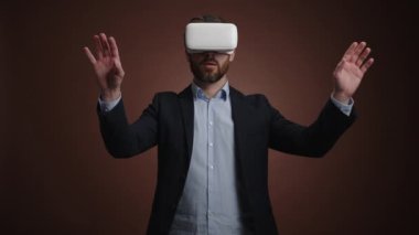 Takım elbiseli ve VR kulaklıklı bir adam bir şey aşırıyor ve büyütüyor. Sonra da heyecanlanıyor. O, basit bir koyu kahverengi arka plandadır. Şaşırmış görünüyor. Yüksek kalite 4k görüntü