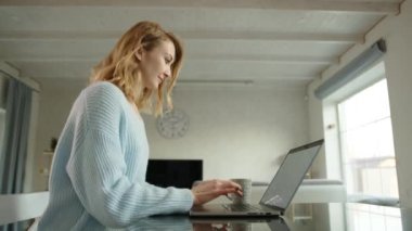 Genç sarışın internette dizüstü bilgisayarından bilgi arıyor. Kadın klavyede yazı yazıyor ve monitöre bakıyor. Arka planda geniş bir oda. Yüksek kalite 4k görüntü