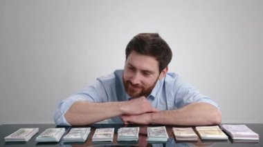 Masada yatan bir adam para yığınlarına bakıyor ve sonra onlardan büyük bir yığın oluşturuyor. Adam açık gri bir odada. Yüksek kalite 4k görüntü