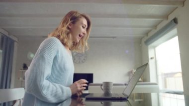 Genç sarışın el sallıyor ve not defteri kullanarak video bağlantısı kuruyor. Kadın ferah bir ışık odasında. Defter, bardak masaya. Yüksek kalite 4k görüntü