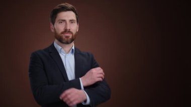 Takım elbiseli bir adam kameraya dönüp gülümseyerek kollarını kavuşturuyor. Güzel giyinmiş ve çok mutlu görünüyor. O, kahverengi bir arka plandadır. Yüksek kalite 4k görüntü