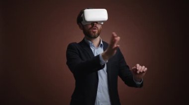 Takım elbiseli ve VR kulaklıklı bir adam bir şeyler aşırıyor ve heyecanlanıyor. O, basit bir koyu kahverengi arka plandadır. Şaşırmış görünüyor. Yüksek kalite 4k görüntü