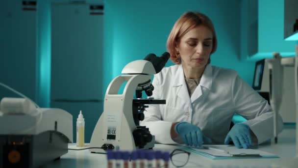 一个穿着白色实验室外套的女人通过显微镜观察样品 写下了一些东西 她在实验室 戴着一副高质的4K胶卷 — 图库视频影像