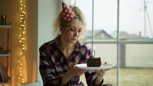 一个戴着生日礼帽的心烦意乱的女人看了看蛋糕 然后看了看窗户 她拿着一个盘子 上面有巧克力蛋糕 高质量的4K镜头 — 图库视频影像