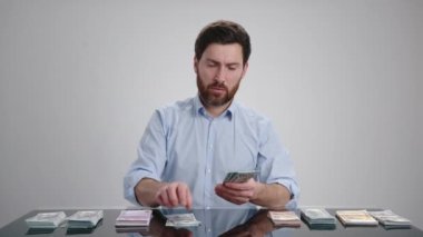 Mavi gömlekli bir adam yeşil para sayıyor ve masaya koyuyor. Odaklanmış görünüyor. Adam açık gri bir odada. Yüksek kalite 4k görüntü 