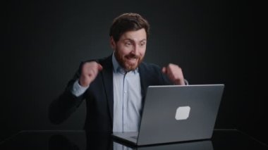 Siyah takım elbiseli bir adam dizüstü bilgisayarda bir şey yazıyor sonra da üstüne para düşerken tezahürat yapıyor. Çok mutlu görünüyor. O, siyah bir odada. Yüksek kalite 4k görüntü