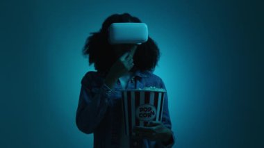 Karanlık odada VR gözlük takan bir kadının portresi. Esmer, Afrika kökenli Amerikalı kız teknoloji programını mavi neon arka plan ve patlamış mısır ile test ediyor. Yüksek kalite 4k görüntü