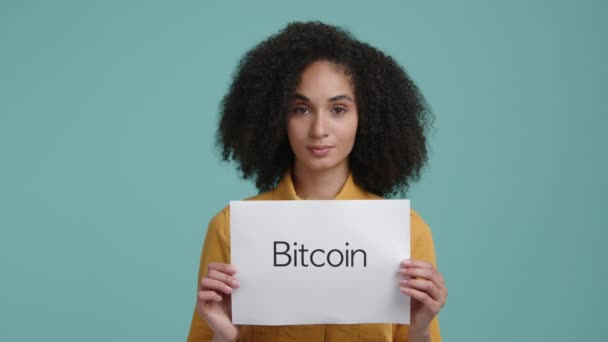 暗号投資を宣伝する少女の肖像画 ビットコインの言葉で白い紙を保持カジュアル服のブルネットの女性 高品質4K映像 — ストック動画