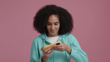 Arkasında pembe bir arka plan olan akıllı telefondan oyun oynayan güzel bir kadın. Aptal ve neşeli görünen genç bir yetişkinin portresi. Yüksek kalite 4k görüntü