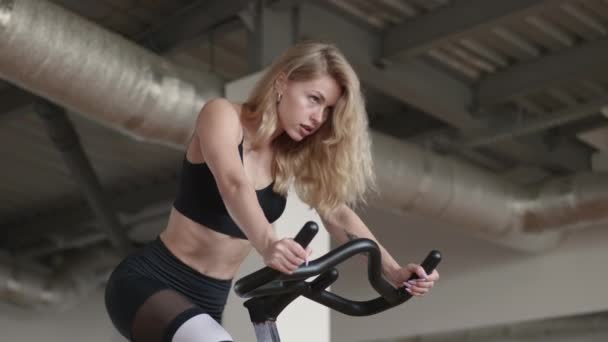 30代の白人の少女は好気性の能力を向上させるためにジムにぶつかった 静止した自転車で運動するかなり運動能力の高い女性 高品質4K映像 — ストック動画