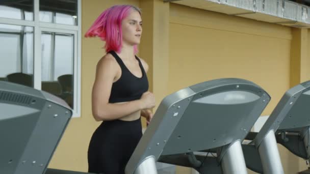 ピンクの髪の美しい若い女性は 減量のためにトレッドミル上で実行しています 健康的でアクティブなスポーツウーマンが屋内でジョギングします 高品質4K映像 — ストック動画