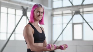 Parlak spor salonunda tek başına antrenman yapan bir kızın portresi. Dambıl egzersizi yaparken gülümseyen neşeli bayan sporcu. Yüksek kalite 4k görüntü