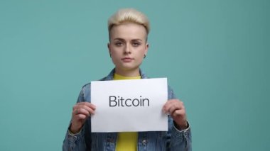 Kripto yatırımı reklamı yapan bir kızın portresi. Üzerinde bitcoin kelimesi olan beyaz bir kağıt tutan günlük giysili beyaz bir kadın. Yüksek kalite 4k görüntü