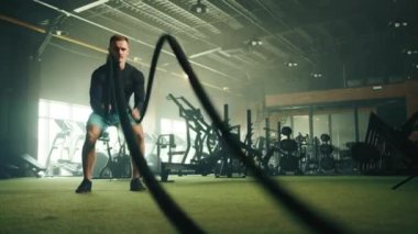 Atletik yakışıklı genç adam tam teçhizatlı spor salonunda savaş halatına vuruyor. Adam siyah uzun kollu sıkıştırma gömleği, spor ayakkabısı ve mavi şort giyiyor. Yüksek kalite 4k görüntü