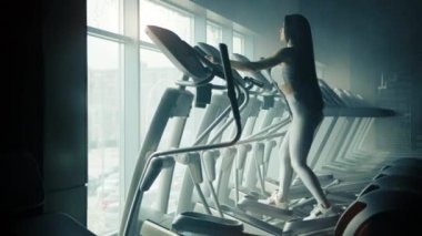 Oldukça yapılı, uzun kahverengi saçlı genç bir kadın eliptik antrenör üzerinde egzersiz yapıyor. Profil fotoğrafı tam uzunlukta. Eliptik spor ayakkabı sırası. Yüksek kalite 4k görüntü