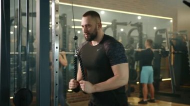 Spor salonundaki dikey sıra makinesine odaklanmış atletik bir gencin yavaş çekim görüntüsü. Erkek ve kadın arka planda halterlerle egzersiz yapıyorlar. Yüksek kalite 4k görüntü