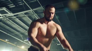 Savaş halatıyla egzersiz yapan çıplak kaslı genç atletik adamın orta derecede yakın çekimi. İp çarpması egzersizleri tek taraflı dalga egzersizleri için değişir. Yüksek kalite 4k görüntü